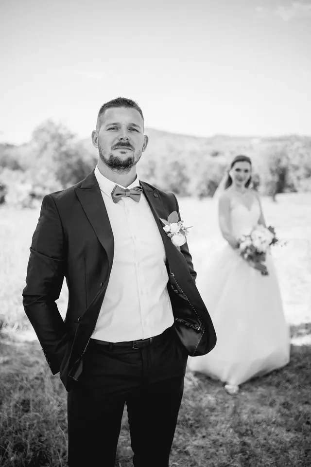 Kitti és Péter: Fekete-fehér vőlegényportré, a menyasszonnyal a háttérben.